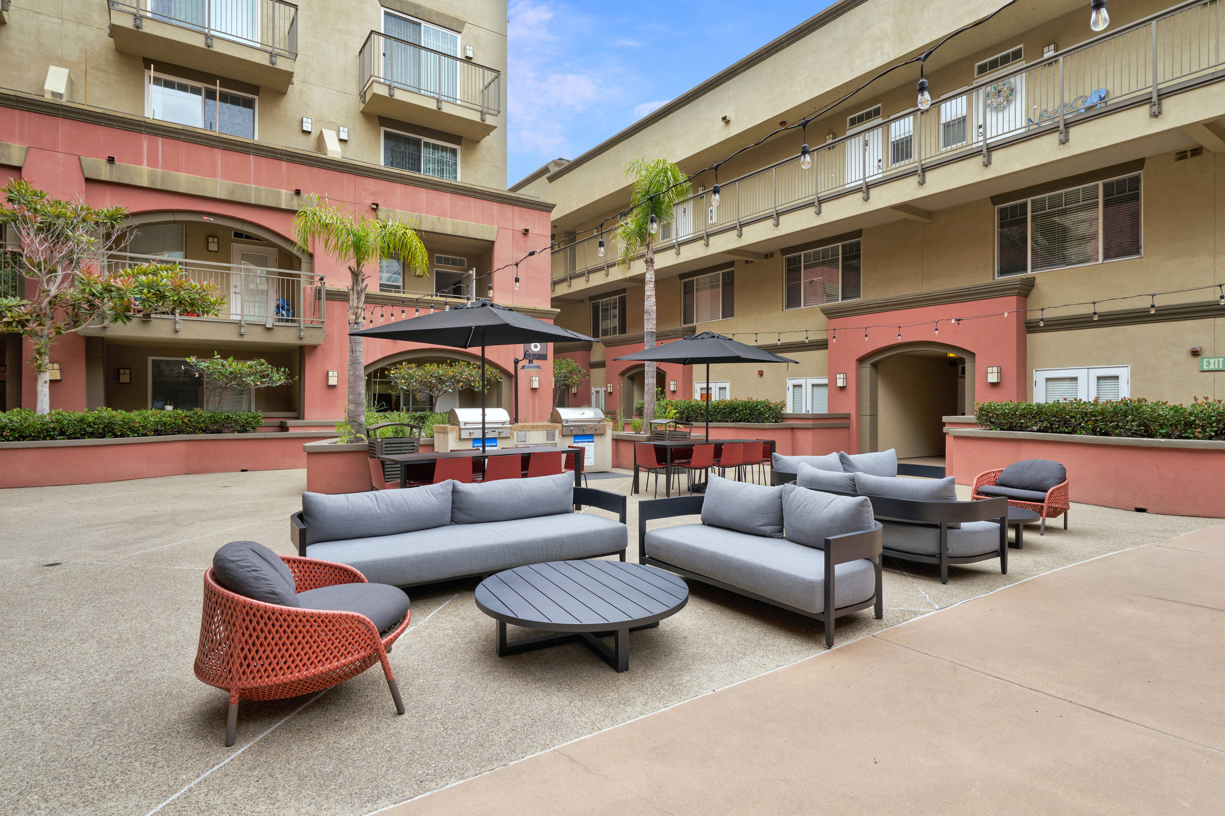 Modern apartment courtyard with outdoor seating, umbrellas, string lights, and palm trees on a sunny day.