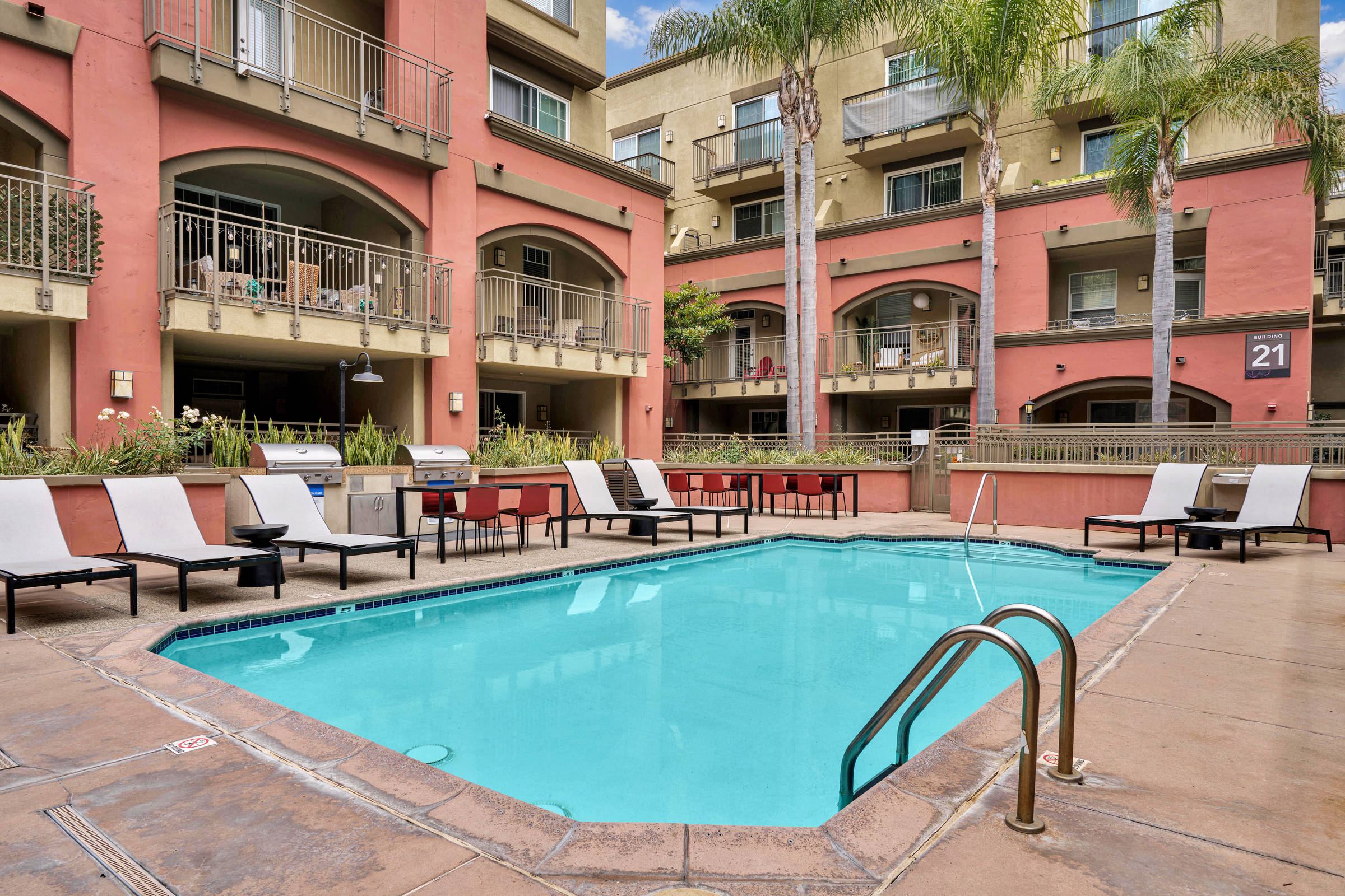 Outdoor apartment pool area with lounge chairs, palm trees, and balconies overlooking the water.