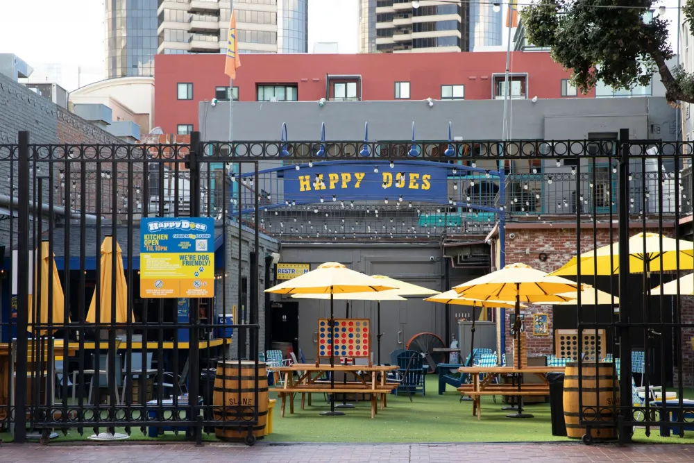 Outdoor patio of a bar called “Happy Does” with yellow umbrellas, string lights, and games, surrounded by tall buildings.
