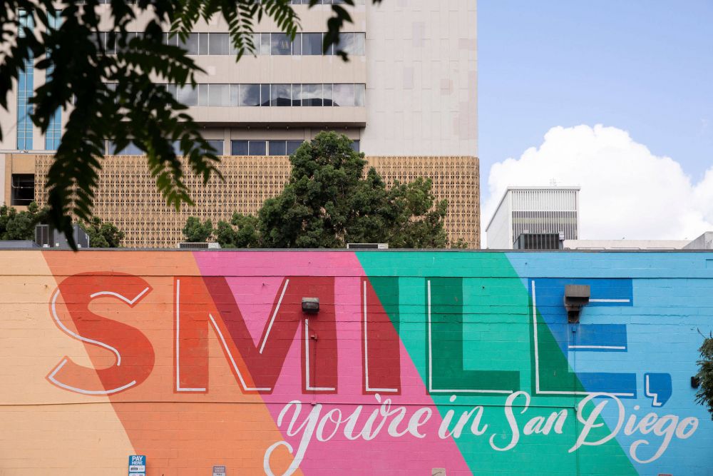 A colorful mural on a brick wall reads “SMILE, you’re in San Diego” in front of a city building and trees.