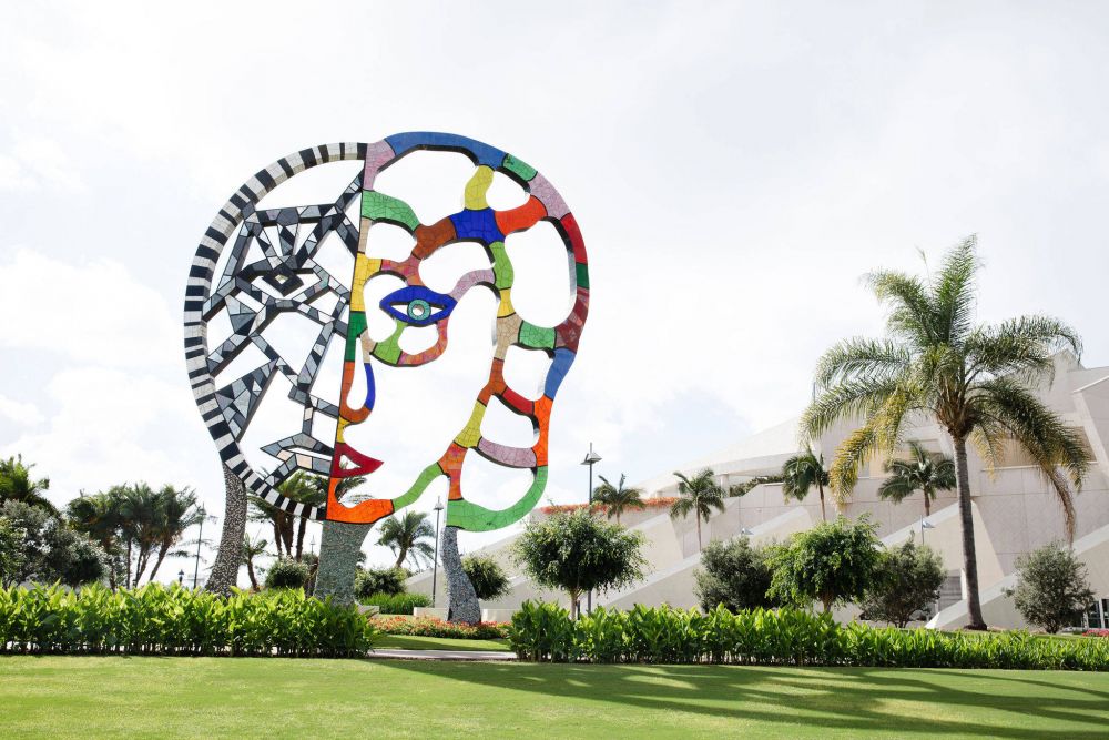 Colorful abstract face sculpture in a garden, with palm trees and a modern building in the background.