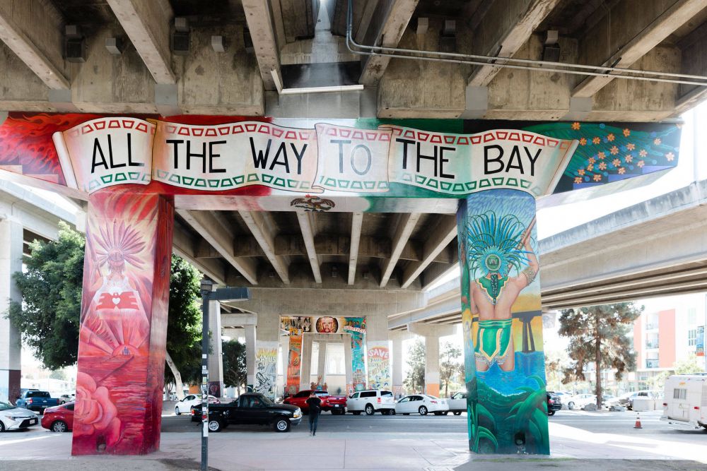 Colorful murals under a highway overpass with a banner reading "ALL THE WAY TO THE BAY" and cars parked below.