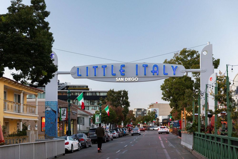 A white "Little Italy San Diego" sign arches over a street lined with cars, trees, and buildings, with Italian flags visible.