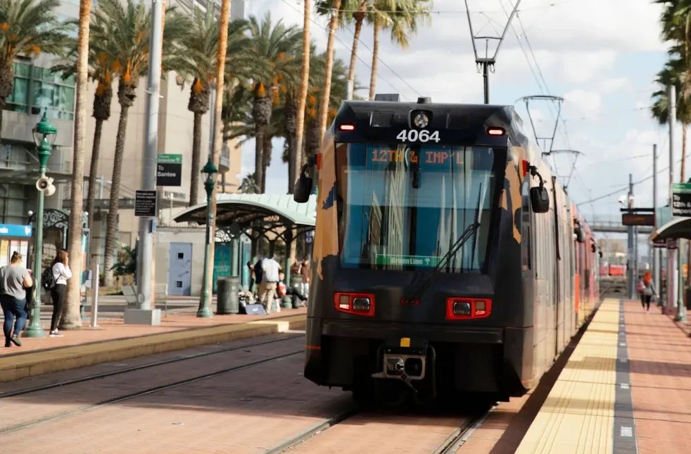 A black trolley train arrives at a palm-lined outdoor station as people wait on the platform.