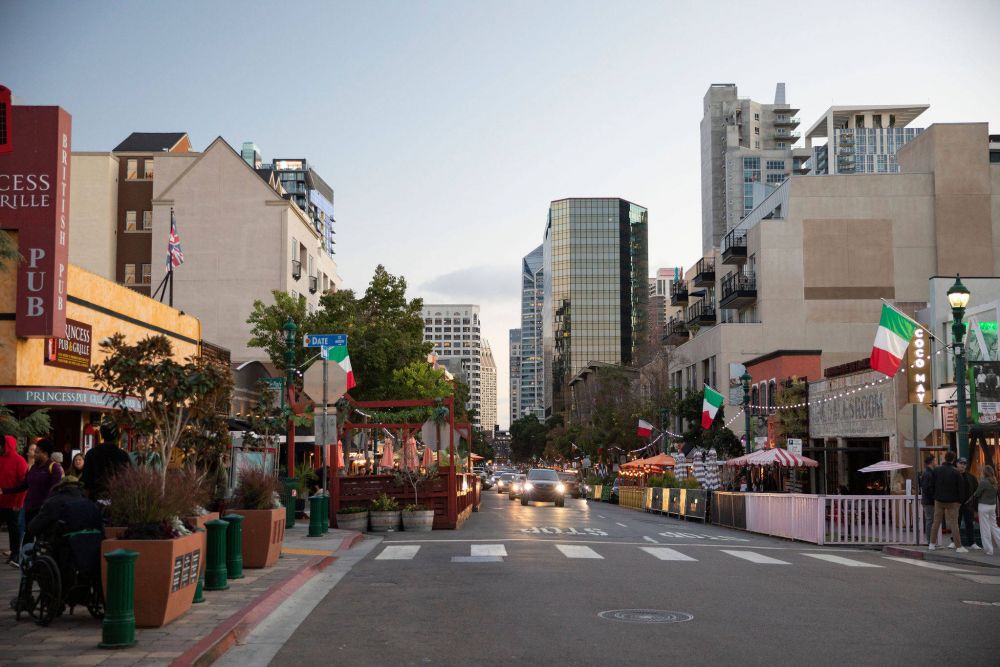 Street scene with outdoor dining, cars, pedestrians, and Italian flags in an urban area with tall buildings in the background.
